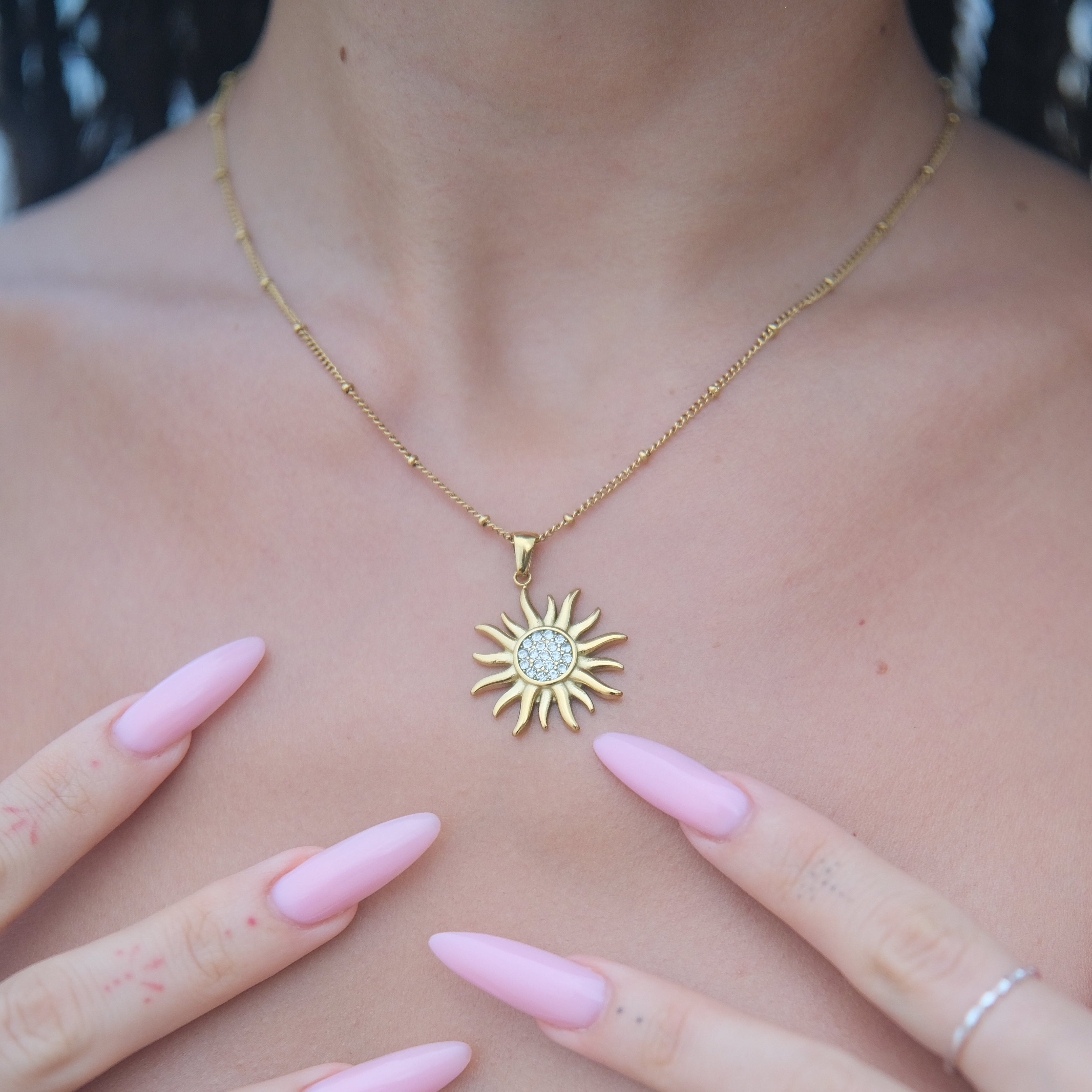 Gold sun-shaped necklace on a person's chest with pink nail polish and rings.